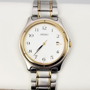 May include: A silver and gold-toned Seiko watch with a white face. The watch has a gold bezel and gold accents on the silver band. The watch face displays the Seiko logo and the date.