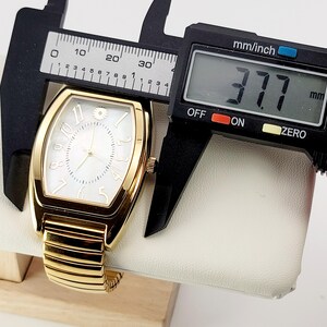 May include: A gold wristwatch with a white face and gold hands. The watch has a rectangular face with a floral accent at the 12 o'clock position. The watch is measured with a digital caliper, showing a width of 37.7 mm.
