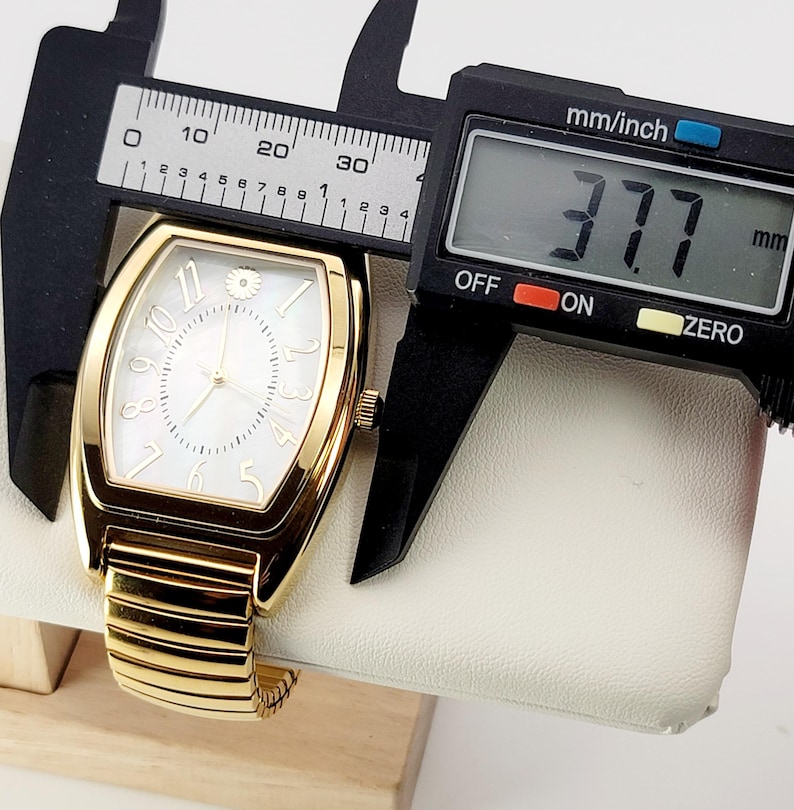 May include: A gold wristwatch with a white face and gold hands. The watch has a rectangular face with a floral accent at the 12 o'clock position. The watch is measured with a digital caliper, showing a width of 37.7 mm.