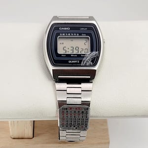 Peut inclure: Montre numérique Casio argentée avec un écran noir affichant l'heure 5:39:20 et la date. La montre a un bracelet argenté et le mot "CASIO" est visible sur le cadran. Un calendrier est visible sur le bracelet.