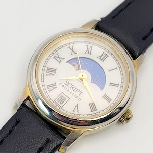 Może przedstawiać: Zegarek na rękę Script Collection z białą tarczą, złotą ramką i czarnym skórzanym paskiem. Tarcza zegarka posiada rzymskie cyfry, wzór fazy księżyca i datę. Zegarek ma złote wskazówki i złotą koronę.