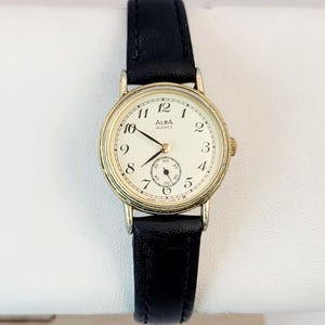 May include: A gold-toned Alba Quartz wristwatch with a cream-colored face and black leather band. The watch features black numerals, a small seconds dial, and the brand name "ALBA QUARTZ".