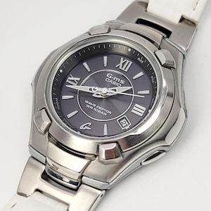 May include: A silver G-MS Casio Wave Ceptor watch with a dark gray face and white leather band. The watch features Roman numeral hour markers and a date display. The band has a textured, crocodile-like pattern.