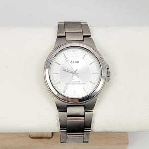 May include: A silver-toned Alba watch with a round face and a matching metal band. The watch face is light-colored with the brand name "ALBA" at the top. The watch is displayed on a white surface.