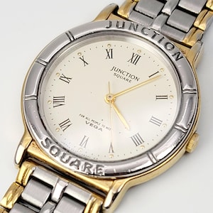 May include: A gold and silver-toned wristwatch with a cream-colored face. The watch features Roman numeral hour markers and the words "JUNCTION SQUARE" on the face and bezel. The watch band is a combination of gold and silver links.