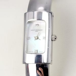 May include: A silver Saint Honore Paris watch with a rectangular face and a mother-of-pearl dial. The watch features a silver band and is adorned with small diamond accents. The watch has the words "Swiss Made" on the face.