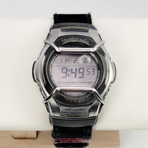 Op de afbeelding: Een zilveren en zwarte Baby-G digitale horloge met de tijd 9:49:53. Het horloge heeft een zwarte band en toont de tekst "WATER 10 BAR RESIST" en "SHOCK RESIST". Het horloge ligt op een lichtgekleurd oppervlak.