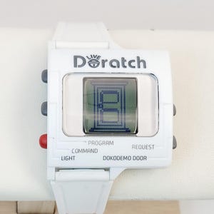 Op de afbeelding: Witte digitale horloge met de tekst "LIVE Doratch" en een scherm met een blauwe afbeelding. Het horloge heeft knoppen met de labels "PROGRAM", "COMMAND", "REQUEST", "LIGHT" en "DOKODEMO DOOR". Het horloge ligt op een wit oppervlak.