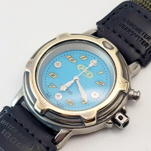 May include: A Fossil Industry watch with a light blue face, featuring gold numerals and hands. The watch has a silver-tone case and a black leather strap. The watch face includes the text "Fossil" and "Meters". The strap has the text "Thermometer Altimeter".