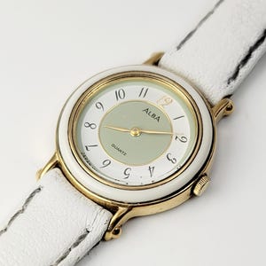 May include: A gold-toned ALBA Quartz watch with a white face and a light green center. The watch has gold hands and a white leather strap. The watch face displays the numbers 1 through 12.