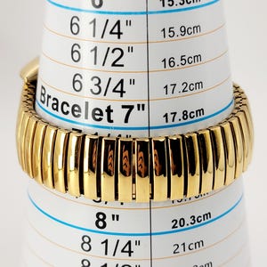May include: A gold-coloured bracelet, approximately 7 inches (17.8 cm) long, is shown on a white measuring tool. The bracelet features a ribbed design and is measured against a scale indicating bracelet sizes in inches and centimetres.