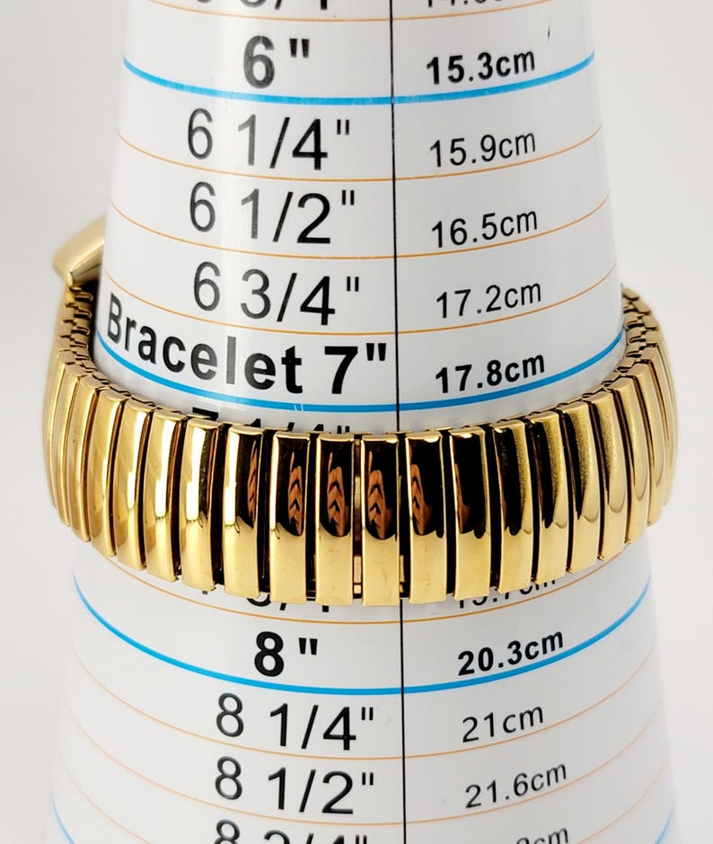 May include: A gold-coloured bracelet, approximately 7 inches (17.8 cm) long, is shown on a white measuring tool. The bracelet features a ribbed design and is measured against a scale indicating bracelet sizes in inches and centimetres.
