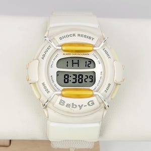 Op de afbeelding: Witte Baby-G digitale horloge met een witte band. De wijzerplaat geeft de tijd en datum weer. Kenmerken zijn schokbestendigheid en een alarmchronograaf. Het horloge heeft gouden accenten en is ontworpen voor dagelijks gebruik.