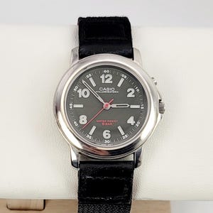 Op de afbeelding: Een Casio Illuminator horloge met een zwarte band. Het horloge heeft een zilverkleurige kast en een donkergrijze wijzerplaat met witte cijfers en wijzers. De wijzerplaat bevat ook de tekst "WATER RESIST 5 BAR".