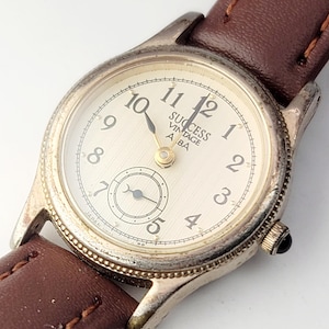May include: A wristwatch with a silver-tone metal case, a cream-coloured dial featuring black Arabic numerals, and the text SUCCESS VINTAGE ALBA. It includes a small sub-dial at the six o'clock position and a brown leather strap with stitching.