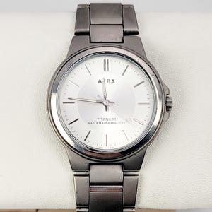 May include: A silver-toned wristwatch with a light-colored face and a matching metal band. The watch face displays the brand name "ABA" and the text "TITANIUM WATER 10 BAR RESIST". The watch is displayed on a white surface.