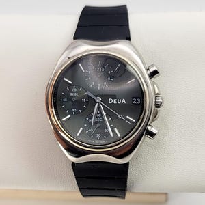 May include: A silver-toned DEUA wristwatch with a black face and a black rubber strap. The watch features multiple sub-dials, including a minute counter and a second counter. The date is displayed at the 3 o'clock position.