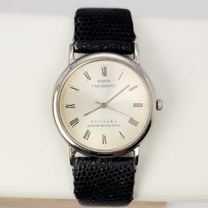 Könnte beinhalten: Silberne Armbanduhr mit einem schwarzen, strukturierten Lederarmband. Das silberne Zifferblatt hat schwarze römische Ziffern und den Text "NIHON UNIVERSITY FUJISAWA SENIOR HIGH SCHOOL".