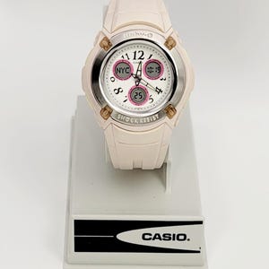 Op de afbeelding: Een lichtroze Baby-G horloge met een witte wijzerplaat en zilveren rand. Het horloge geeft de tijd en datum weer, met de tekst "NYC" en "SHOCK RESIST" zichtbaar. Het horloge staat op een witte standaard met de merknaam "CASIO."