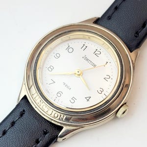 May include: A silver-toned analog watch with a white face, gold hands, and black leather band. The watch face displays the word "Junction" and the brand name "VEGA". The watch has a round face and a black leather strap.