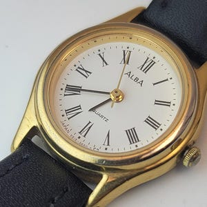 May include: Gold-toned Alba quartz watch with a white face and black Roman numerals. The watch has a black leather band and a gold bezel. The watch face displays the word "ALBA" and "QUARTZ". The watch is a classic timepiece.