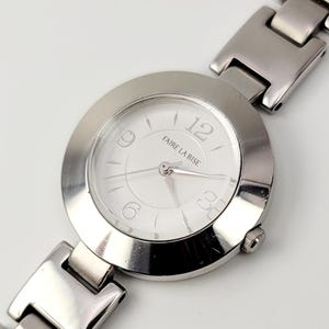 Puede incluir: Un reloj de pulsera plateado con esfera blanca. El reloj presenta el texto "FAIRE LA BISE" y tiene una correa de metal. La esfera muestra los números 12, 3, 6 y 9.
