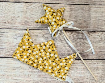 May include: A yellow crown and star wand with white floral print. The crown has a white ribbon tie and the wand has a white ribbon and wooden stick.