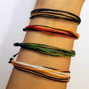 May include: A stack of colorful friendship bracelets on a person's wrist. The bracelets are made of thin, braided cords in black, white, orange, green, red, and tan.