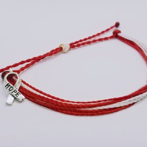 May include: Red, white, and silver bracelet with a ribbon charm that says "HOPE". The bracelet is made of woven red and white cords. The charm is a silver ribbon. The bracelet is a symbol of support and awareness.