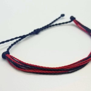 Puede incluir: Pulsera hecha a mano con cordones rojos y azul marino. La pulsera está hecha de múltiples hebras, anudadas en los extremos. La combinación de colores es un contraste audaz, adecuado para el uso diario. Aproximadamente 15-20 cm de largo.