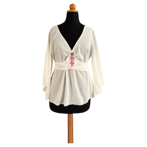 Könnte beinhalten: Weiße, halbtransparente Bluse mit langen Ärmeln, V-Ausschnitt und einem pinkfarbenen, floralen Stickdetail in der Mitte vorne. Die Bluse hat eine geraffte Taille und eine leicht ausgestellte Silhouette.