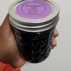 May include: A jar of blueberry jam with a silver lid and a purple label. The label reads "Locked in the Garden Blueberry Jam." The jar is held in a hand and has a diamond pattern.