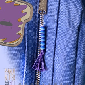 May include: A close-up of a blue zipper pull with a purple tassel and a blue and white beaded charm. The zipper pull is attached to a blue fabric background with the word "HERSCHEL" printed on the zipper.