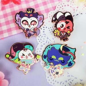 Puede incluir: Cuatro pines de esmalte de colores con personajes de dibujos animados con diferentes expresiones. Los pines están sobre un fondo a cuadros rosa y blanco con doilies blancos.