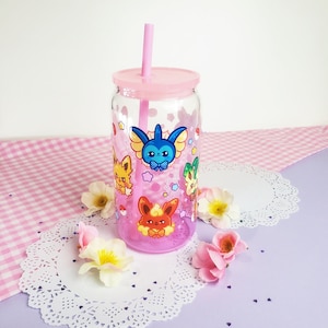 May include: A pink glass tumbler with a pink lid and a pink straw. The tumbler has a clear glass body with a design of cartoon characters from the Pokemon franchise. The characters are surrounded by stars and hearts.