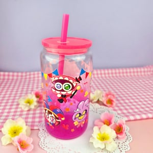 Puede incluir: Un vaso de plástico transparente con tapa y pajita rosas, lleno de líquido rosa. El vaso está decorado con personajes de dibujos animados y banderas de colores. El vaso está sobre un tapete blanco con flores rosas y amarillas.