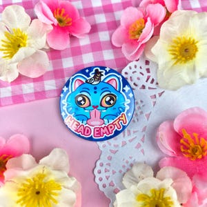 May include: A round, blue button with a cartoon cat face that has large eyes and a long tongue. The button has the words "HEAD EMPTY" in a red font. The button is surrounded by pink and white flowers.