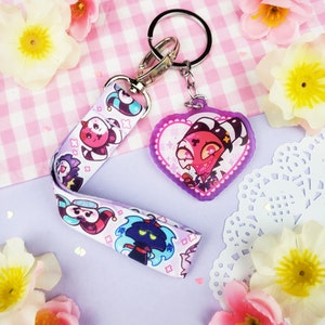 May include: A pink and white heart-shaped keyring with a cartoon character with red horns and a black and white striped shirt. The keyring is attached to a white and pink lanyard with cartoon characters.