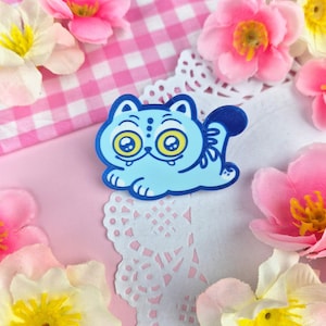 May include: A light blue cat-shaped enamel pin with large yellow eyes and a dark blue tail. The pin is surrounded by pink and yellow flowers and rests on a pink and white checkered background.