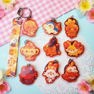 May include: A selection of vibrant acrylic keyrings showcasing cartoon characters. The keyrings are predominantly orange, red, and yellow, with silver key rings. A coordinating lanyard with cartoon character designs is also present.