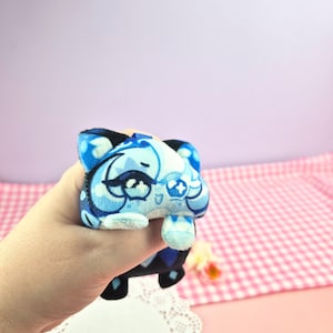 May include: A small, plush toy in the shape of a cartoon character, primarily in shades of blue and white. The toy has a detailed face and is held in a hand. The background is a light pink and white checkered pattern.