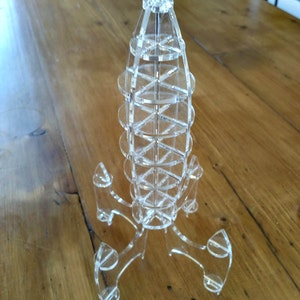 Op de afbeelding: Een helder acrylmodel van een raket met een puntige top en een basis met drie poten. Het model is gemaakt van meerdere lagen acryl die aan elkaar zijn verbonden.