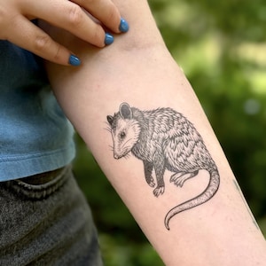 May include: A black and white tattoo on a forearm featuring a detailed illustration of an opossum. The animal is depicted in profile with fine line work showing its fur texture, pointed snout, small ears, and a long, curved, hairless tail.