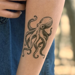 Könnte beinhalten: Ein temporäres Tattoo mit einem detaillierten, schwarz-weißen Tintenfisch-Design auf dem Unterarm. Das Tattoo zeigt die komplexen Details des Tintenfischs. Das Tattoo befindet sich auf einem Arm, vor einem Jeanshemd-Hintergrund.