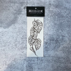 May include: Black and white temporary tattoo featuring a design of a Monstera plant with leaves.