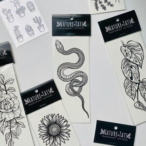 May include: A set of temporary tattoos featuring black and white illustrations of a snake, sunflower, monstera leaf, and a rose. The tattoos are packaged on individual sheets with the brand name "NATURE-TATS" and the words "TEMPORARY TATTOOS".