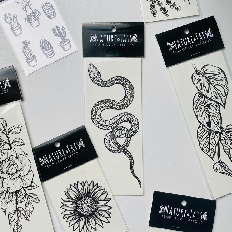 May include: A set of temporary tattoos featuring black and white illustrations of a snake, sunflower, monstera leaf, and a rose. The tattoos are packaged on individual sheets with the brand name "NATURE-TATS" and the words "TEMPORARY TATTOOS".