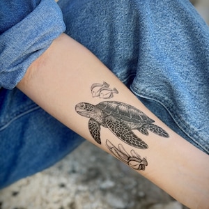 May include: Black and white temporary tattoo of a sea turtle, two fish, and a sea plant. The tattoo is on a person's arm, with blue denim jeans in the background. The sea turtle is the focal point of the tattoo.