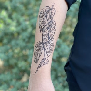 May include: A black and white temporary tattoo of a monstera leaf design on a person's arm.
