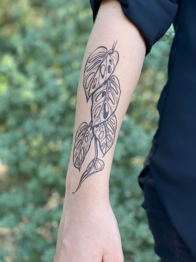 May include: A black and white temporary tattoo of a monstera leaf design on a person's arm.